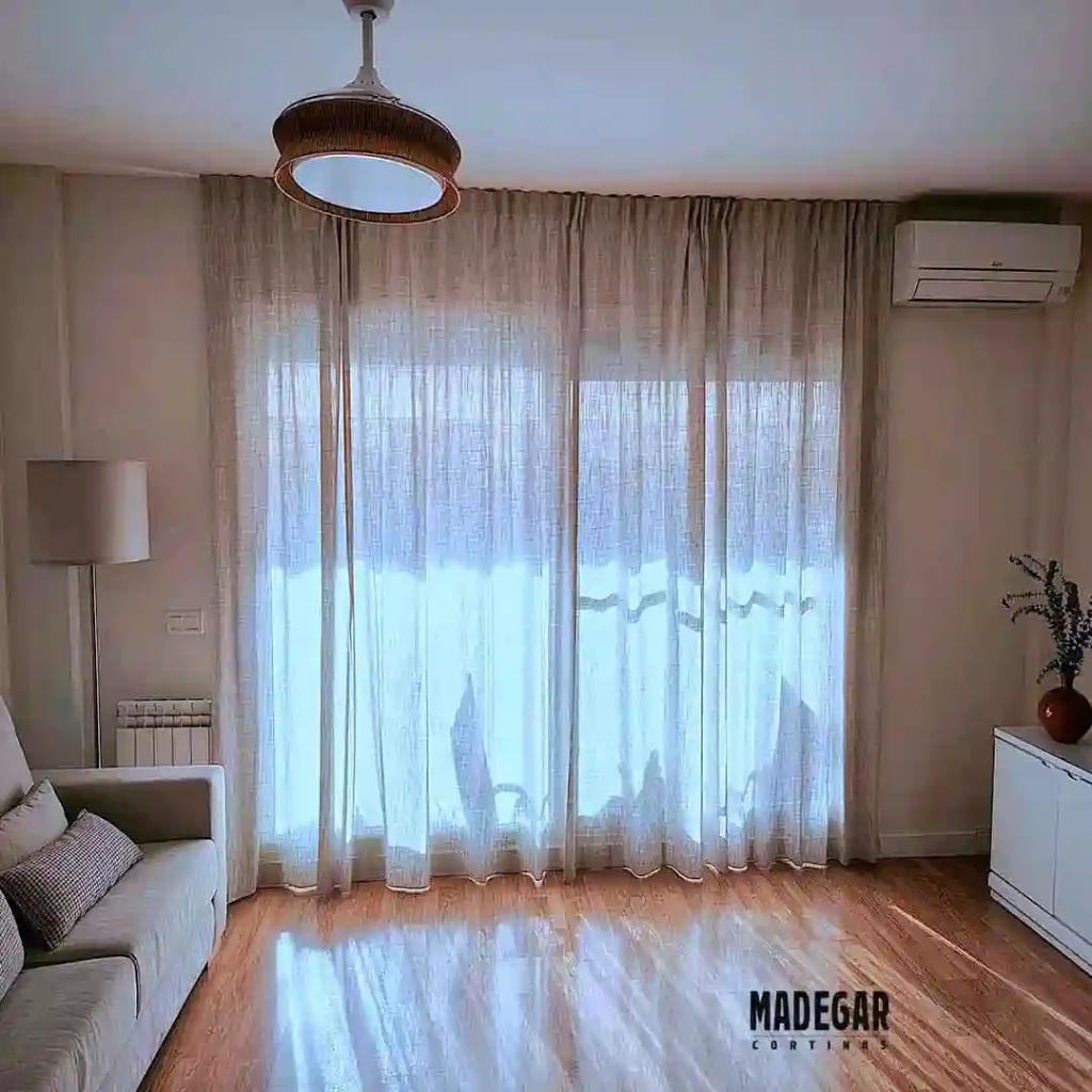 Cortinas convencionales confeccionadas a medida en tejido de semi-lino natural de estilo informal instaladas en el ventanal del salón de un piso de alquiler.