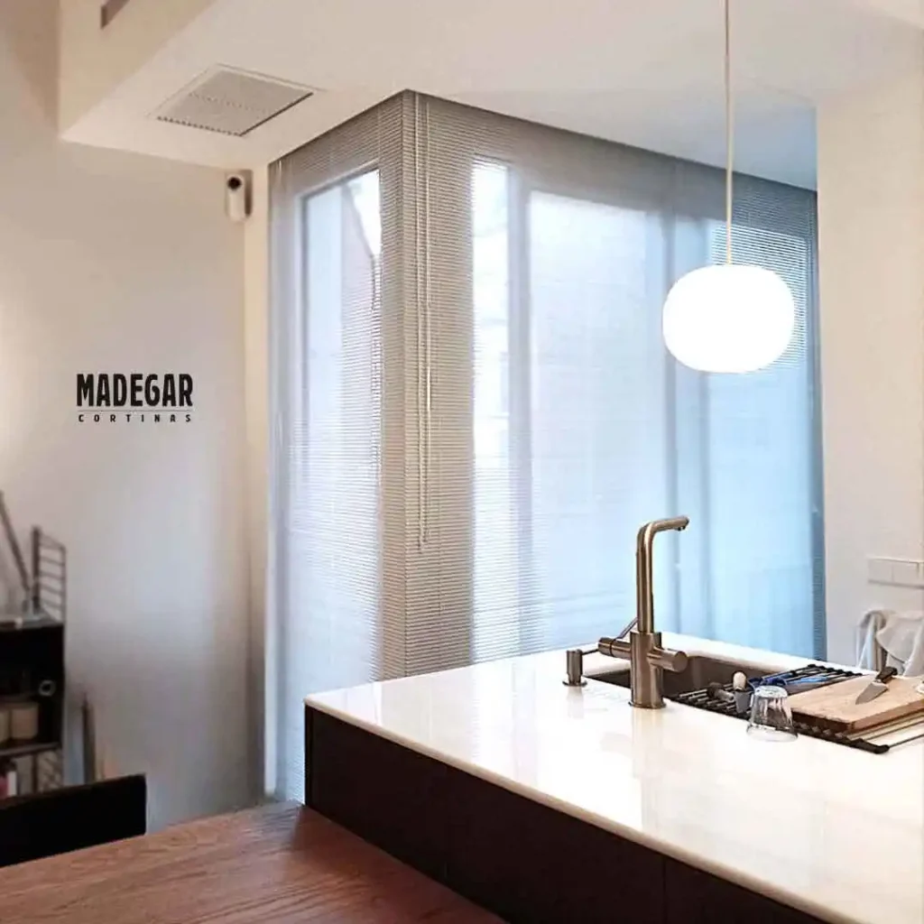 Cortina veneciana de aluminio blanco con lamas compactas de 16 mm instalada por Madegar Cortinas en la tribuna acristalada de una cocina de diseño abierta, regulando de forma milimétrica la luz natural y la intimidad frente al patio interior de manzana del Eixample.