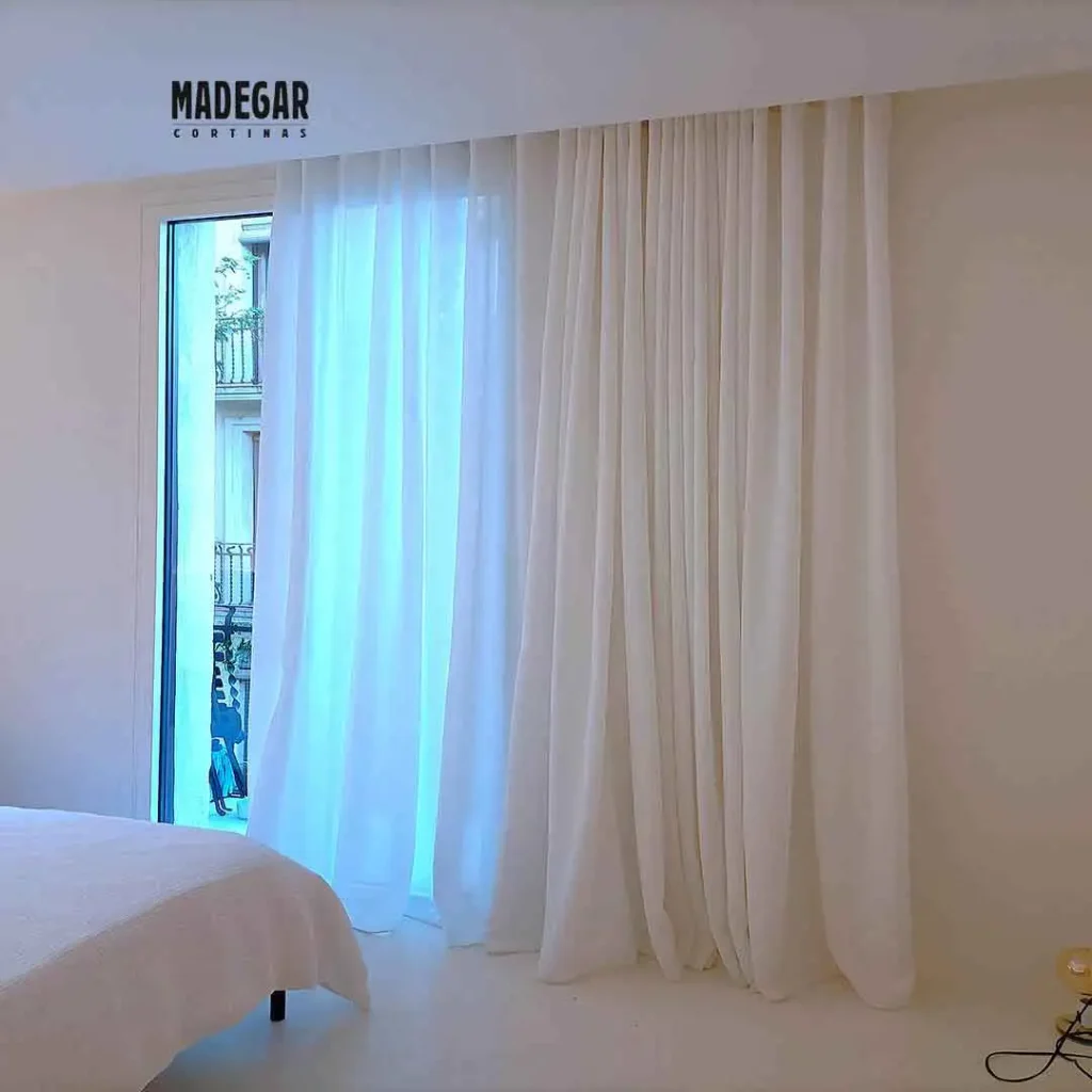 Cortinas a medida para el dormitorio de un loft del Eixample instaladas por Madegar Cortinas, confeccionadas en tela blanca de lino con sistema de onda perfecta y caídas generosas que arrastran sutilmente sobre el suelo claro, vistiendo la salida al balcón y regulando la entrada de luz natural en un piso del Eixample Barcelona.