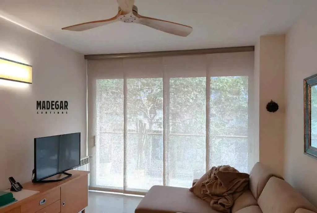 Panel japonés de 4 vías confeccionado a medida en tejido screen translúcido, instalado por Cortinas Madegar con un perfil embellecedor superior que oculta el mecanismo del riel en la salida al balcón de un salón en el Eixample de Barcelona