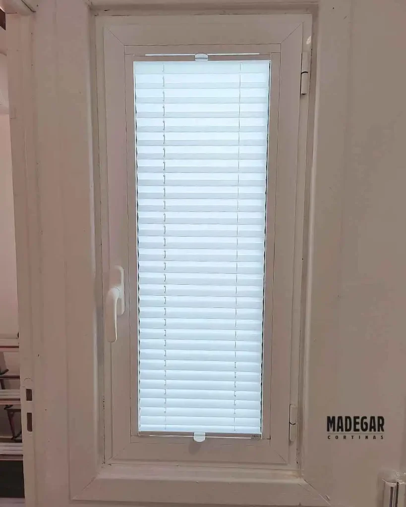 Sistema plisado compacto fijado directamente al junquillo de una ventana oscilobatiente de aluminio blanco.