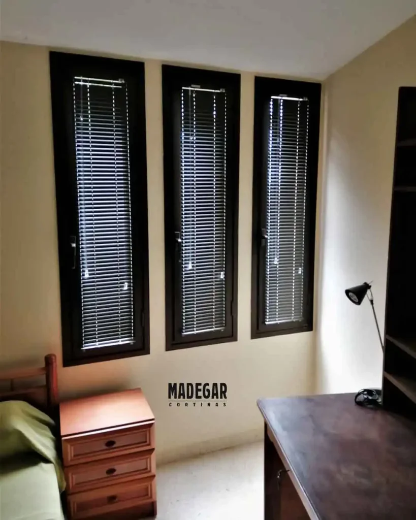 Venecianas técnicas con lamas de aluminio estrechas instaladas directamente sobre los marcos de tres ventanas oscilobatientes consecutivas.