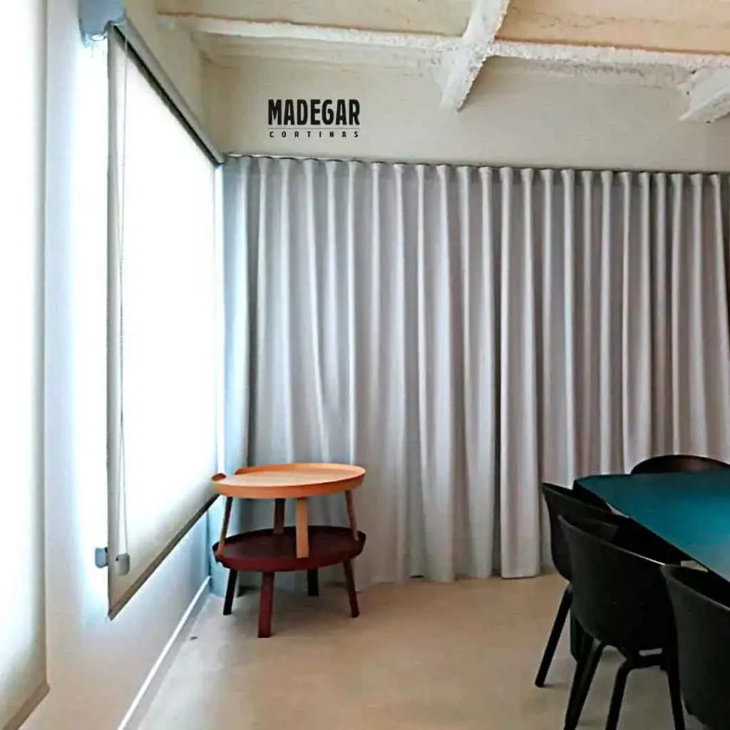 Separación de espacios de trabajo mediante cortinas técnicas fonoabsorbentes en gris claro y estor enrollable de screen en ventana exterior, ideal para mitigar el ruido y los reflejos en oficinas.