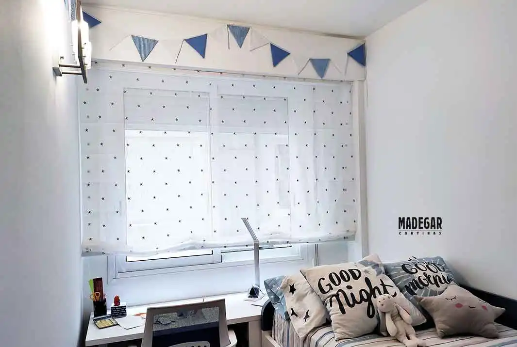 Detalle de una acogedora habitación infantil en Sant Cugat del Vallès, Barcelona. Un estor paqueto de tela blanca con estampados de estrellas cubre el ventanal, tamizando la luz para un ambiente perfecto. Sobre la ventana, un banderín decorativo de tela azul y blanco. La escena incluye una cama con cojines temáticos a juego con la colcha. todo diseñado por Madegar Cortinas 