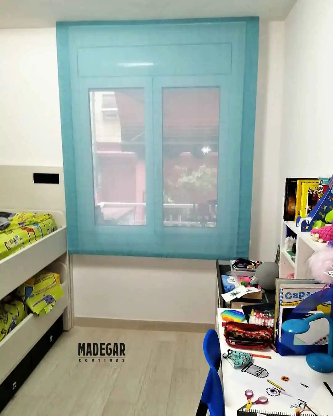Estor paqueto de color turquesa vivo instalado por Madegar Cortinas en un cuarto de niños en el barrio de Gràcia, Barcelona. Se ve cómo queda sobre la ventana, justo al lado de una litera blanca y un escritorio lleno de libros y cosas de clase.
