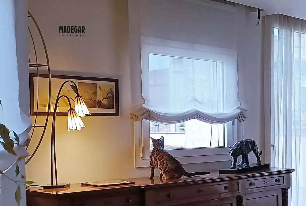 Instalación de estor paqueto a medida en lino blanco para un salón de lujo en el Eixample, Barcelona. Un gato bengalí observa por la ventana sobre un mueble clásico, mostrando la caída natural y elegante de la tela de Madegar Cortinas.