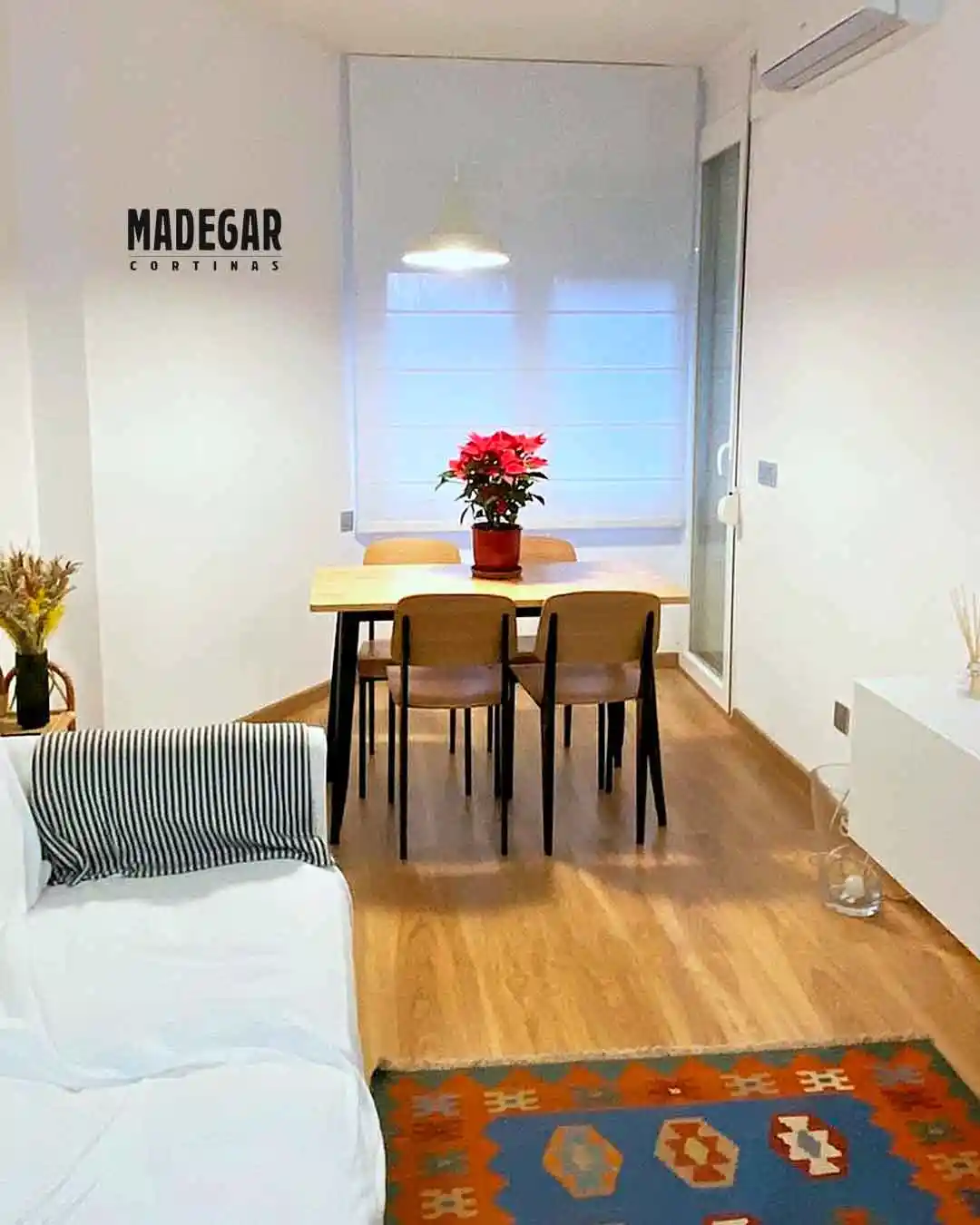 Foto de un comedor pequeñito en un piso de Horta-Guinardó, Barcelona, con una pared que hace una forma irregular. Hemos instalado un estor con varillas de loneta en color natural que queda súper limpio y ordenado sobre la mesa de madera. Se ve el sofá blanco con una mantita a rayas, una alfombra de colores y una planta roja encima de la mesa que le da mucha vida al rincón.