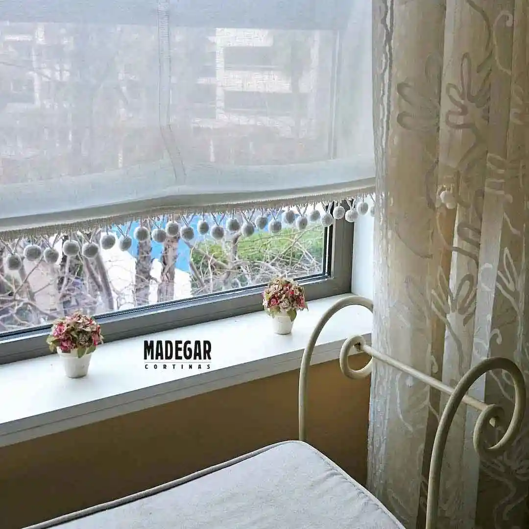 Detalle del acabado inferior de un estor paqueto de tela de lino natural en un dormitorio de una casa en Diagonal Mar. El estor tiene un remate decorativo de pasamanería con pequeñas borlas colgantes de hilo de lino, confeccionado por Madegar Cortinas. En el fondo se ven dos macetas en el alféizar de la ventana y el exterior con árboles.