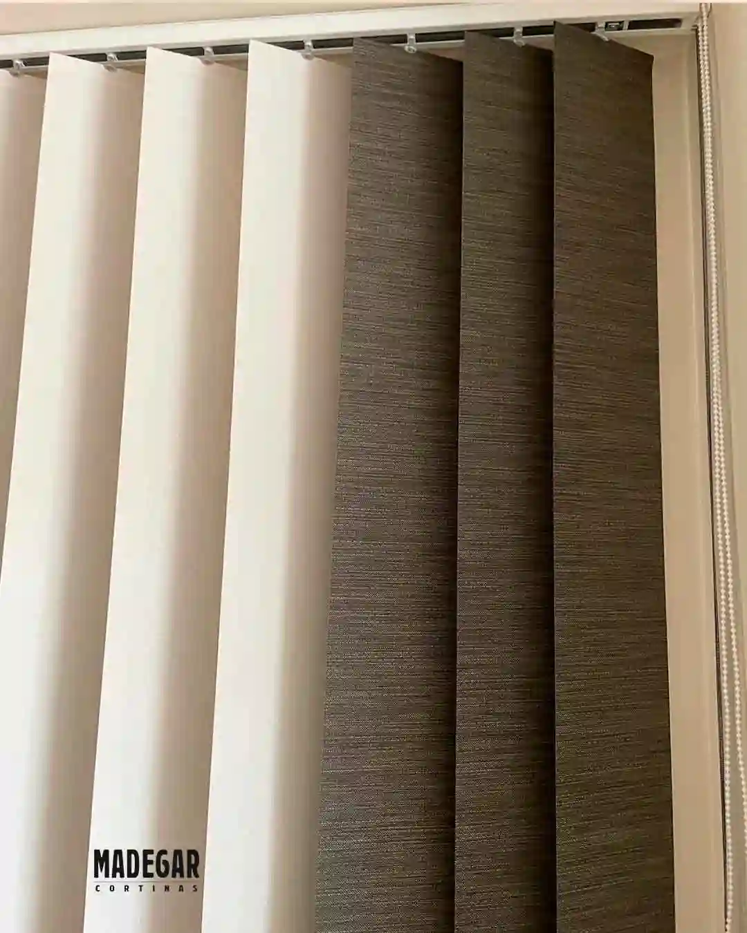 Detalle técnico de una cortina vertical bicolor en tejido screen (blanco y gris antracita) instalada en un salón de Poblenou, Barcelona. Se aprecia el riel superior de aluminio y el sistema de giro de las lamas de Madegar Cortinas.