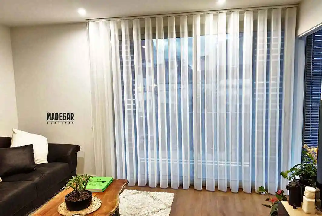 Cortina vertical de tela traslúcida blanca instalada en un salón de dimensiones reducidas en Les Corts, Barcelona. El sistema de Madegar Cortinas permite tamizar la luz natural manteniendo la sensación de amplitud en el espacio.